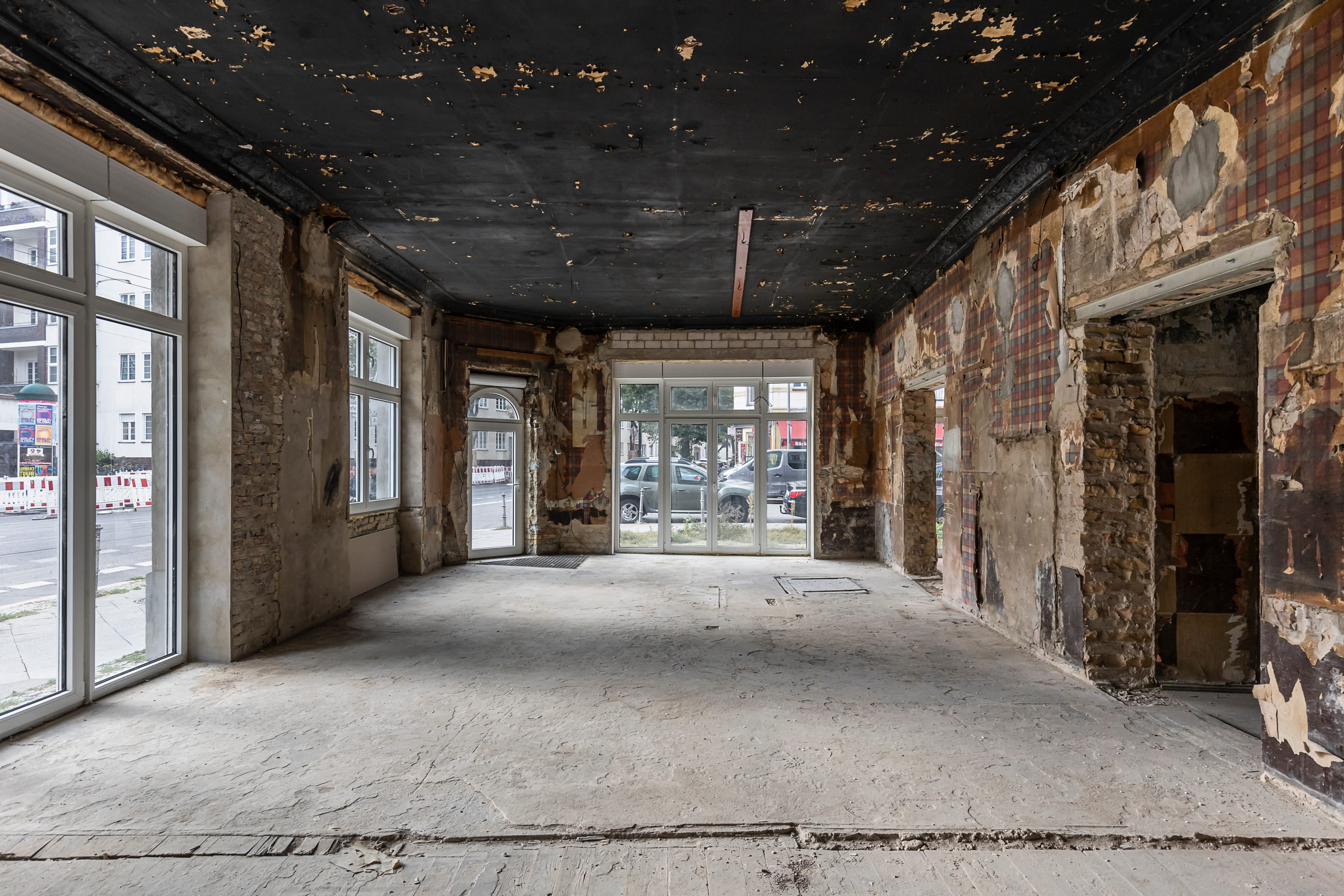 Bestandsaufnahme vor der Sanierung: Eine leere, unfertige Ladenfläche oder ein Loft im Rohbau. Der Innenraum zeigt rohen Betonboden, freiliegende Ziegelwände im Industrielook und eine schwarze, beschädigte Decke. Große, bodentiefe Fenster und Schaufensterfronten zur Straßenseite bieten viel Potenzial für eine moderne Gewerbefläche oder Galerie.